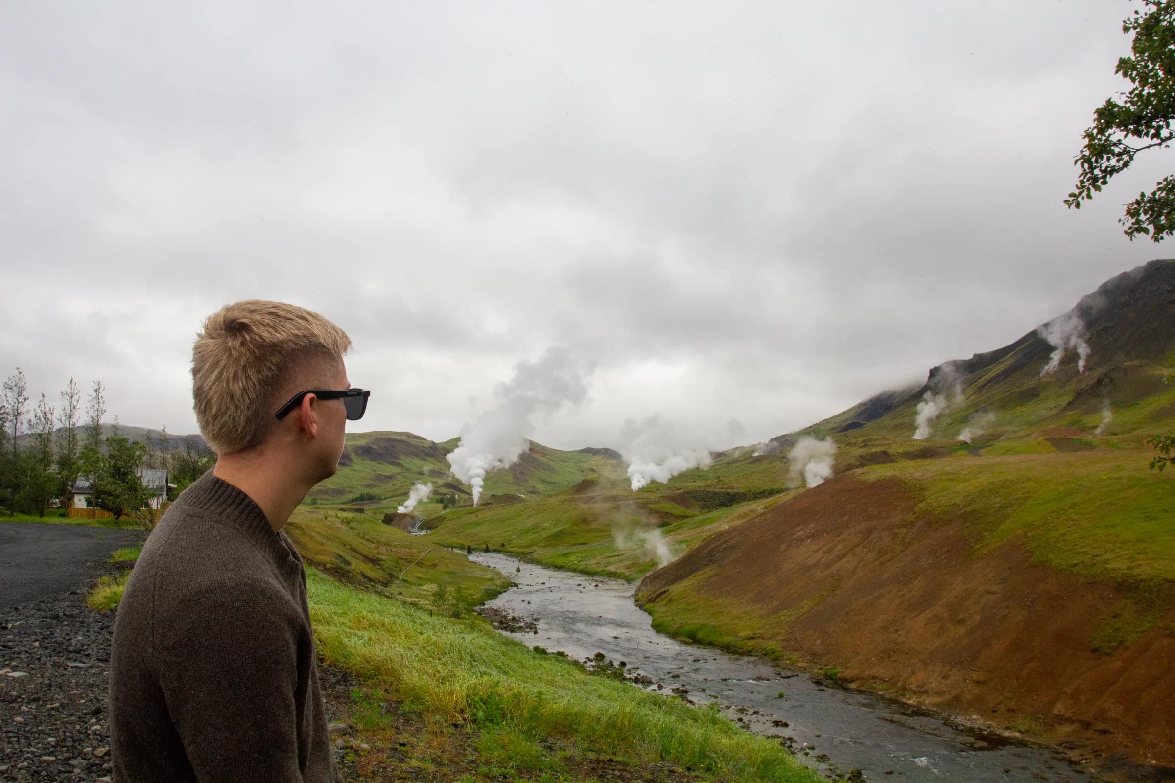 Maður með SonaVue-hljóðgleraugu horfir yfir grænan jarðhitadal á Íslandi, gufa stígur upp úr hverum í hlíðunum.
