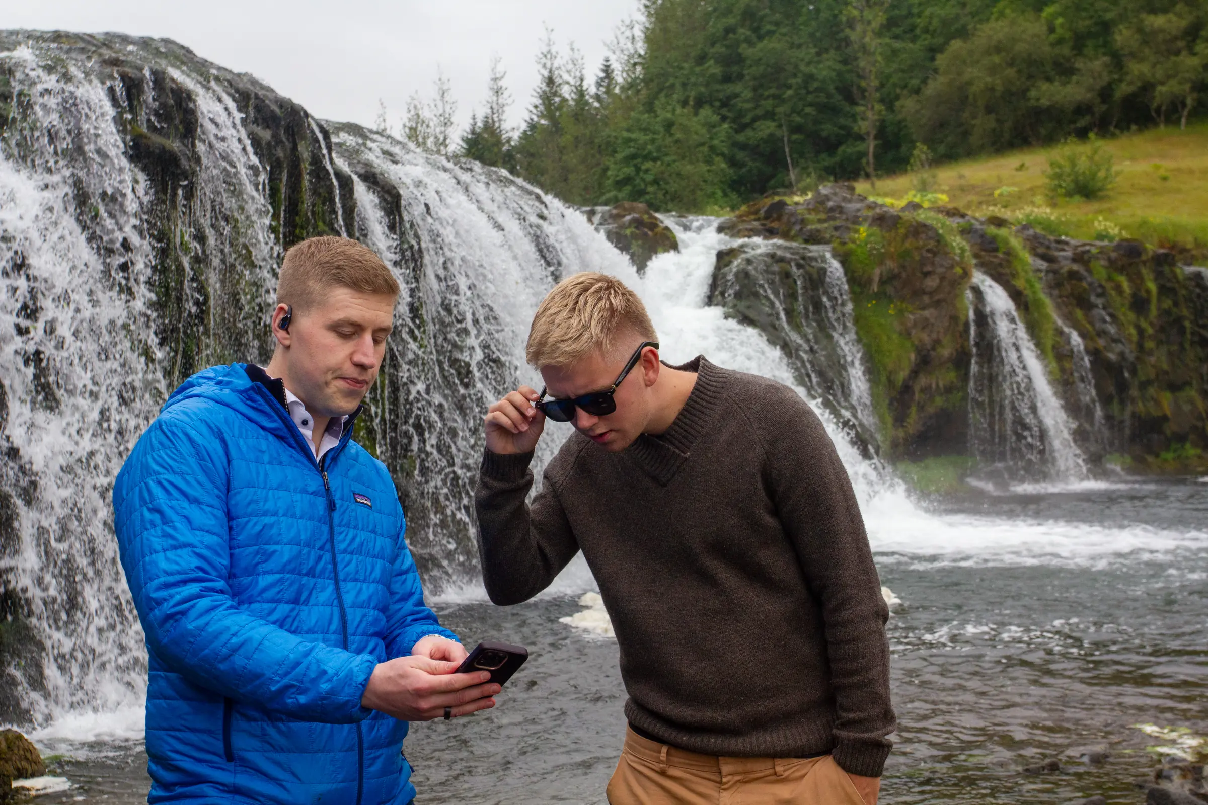 Tveir menn á spjalli við íslenskan foss, annar með TravelerAI-heyrnartól í eyra og hinn snertir spöngina á SonaVue-gleraugunum sínum á meðan þeir skoða appið í síma.